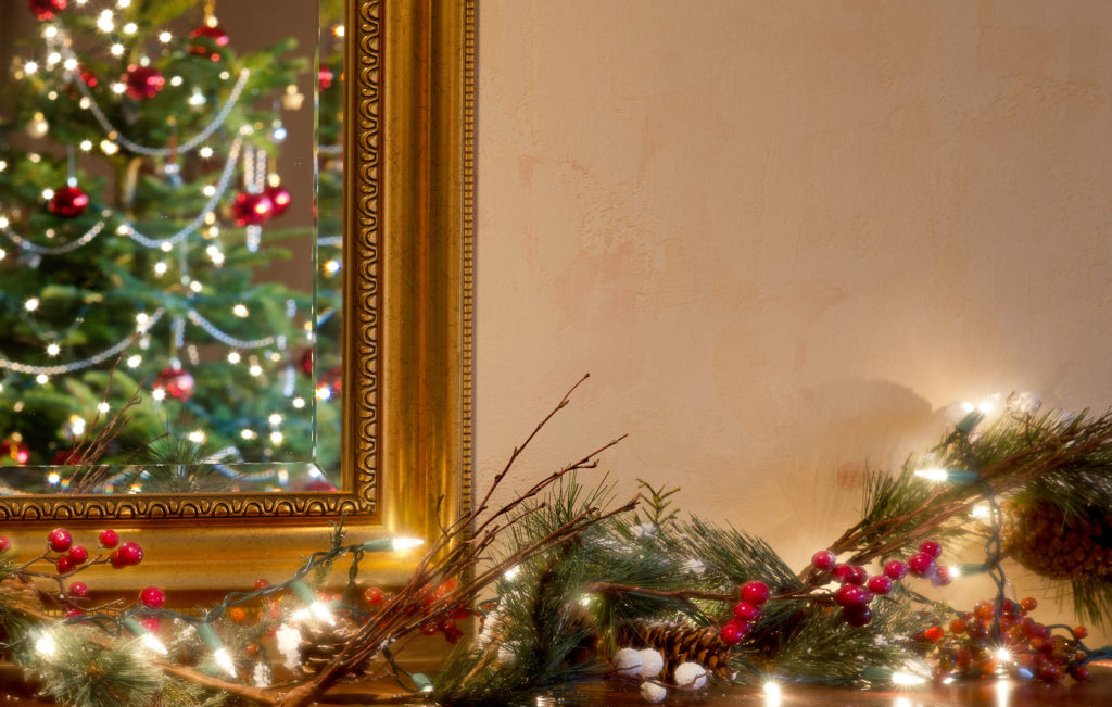 A traditionally decorated Christmas tree is reflected in a mirror over a hearth with a trail of red berries, pine boughs, and twinkling lights. A Tuscan styled wall invites your message.